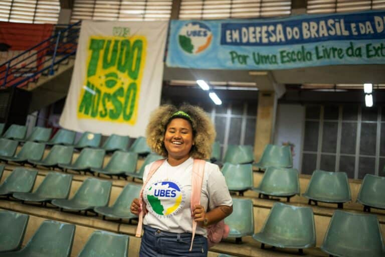 Foto de Roberta Pontes com cartaz da UBES ao fundo e de pé no que parece ser uma arquibancada de ginásio